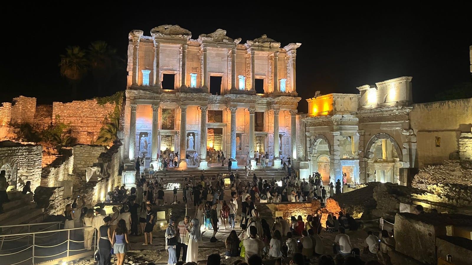 The illuminated Library of Celsus at Ephesus at night, with visitors gathered in front of it.